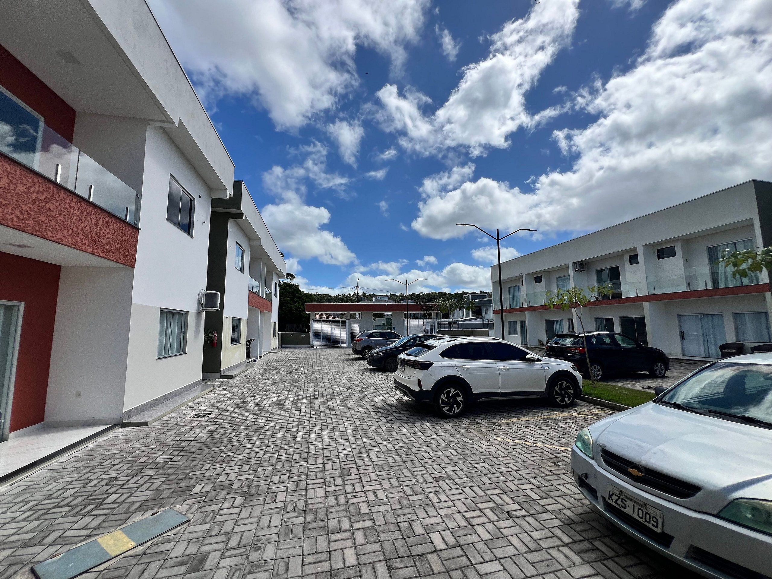 Une aire de stationnement dallée où sont garées plusieurs voitures, un bâtiment moderne doté de balcons et un ciel dégagé avec quelques nuages.