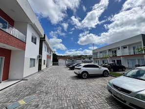 A paved parking area with multiple cars, a modern building with balconies, and a clear sky with clouds.