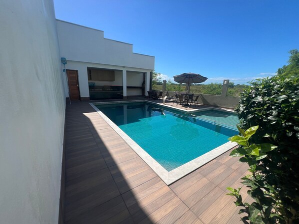 A rectangular swimming pool with a wooden deck surrounding it, a covered seating area with chairs and an umbrella, and a view of greenery in the background.