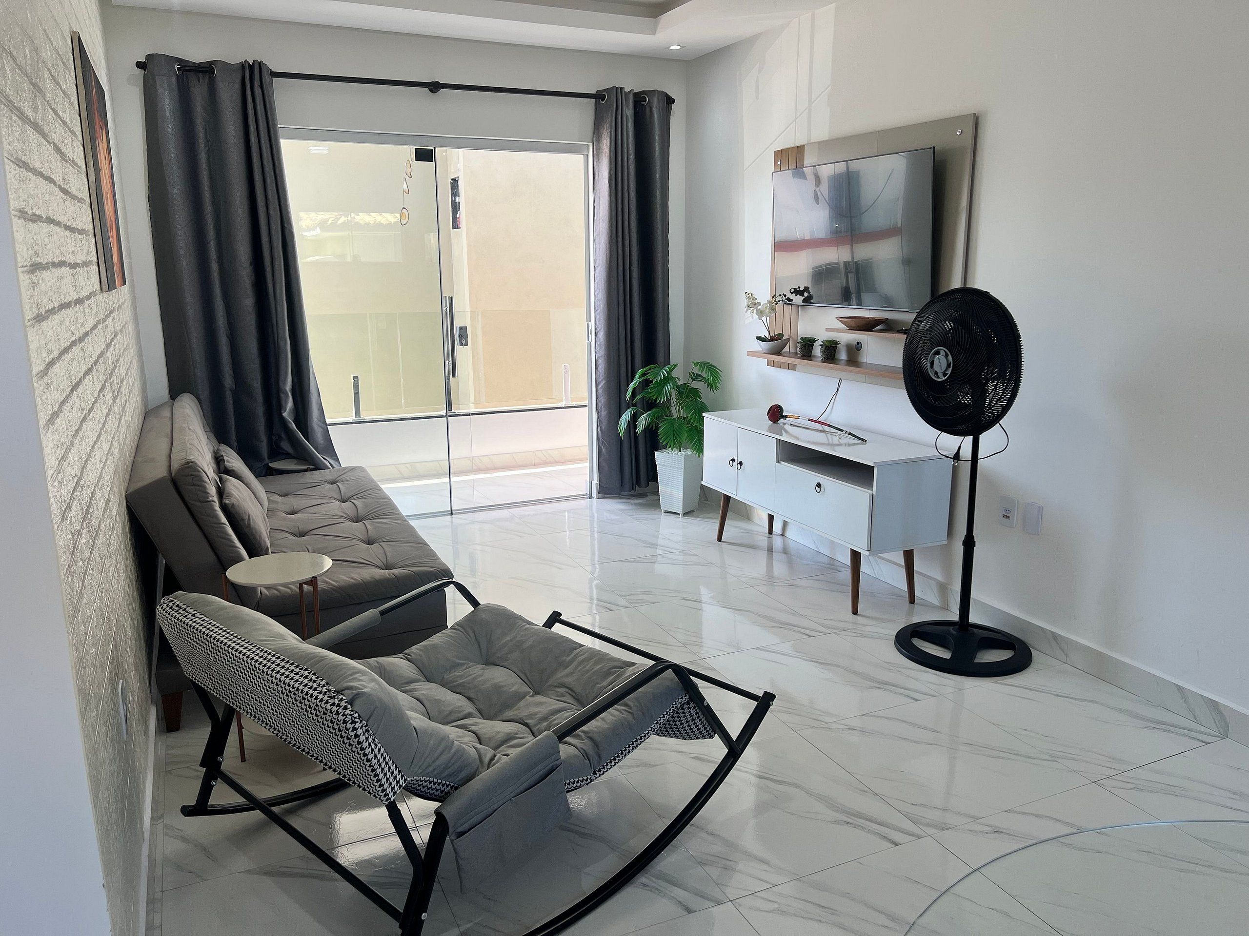 Un salon moderne comprenant un canapé, un fauteuil à bascule, une petite table, un meuble blanc, un ventilateur sur pied et une plante en pot.