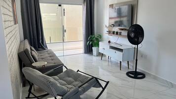 Un salon moderne comprenant un canapé, un fauteuil à bascule, une petite table, un meuble blanc, un ventilateur sur pied et une plante en pot.