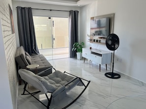 A modern living room with a sofa, a rocking chair, a small table, a white cabinet, a floor fan, and a potted plant.