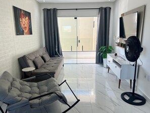 A modern living room with a grey sofa, a small round table, a white cabinet, a large mirror, and a black fan. - Capitania Residence (Porto Seguro)