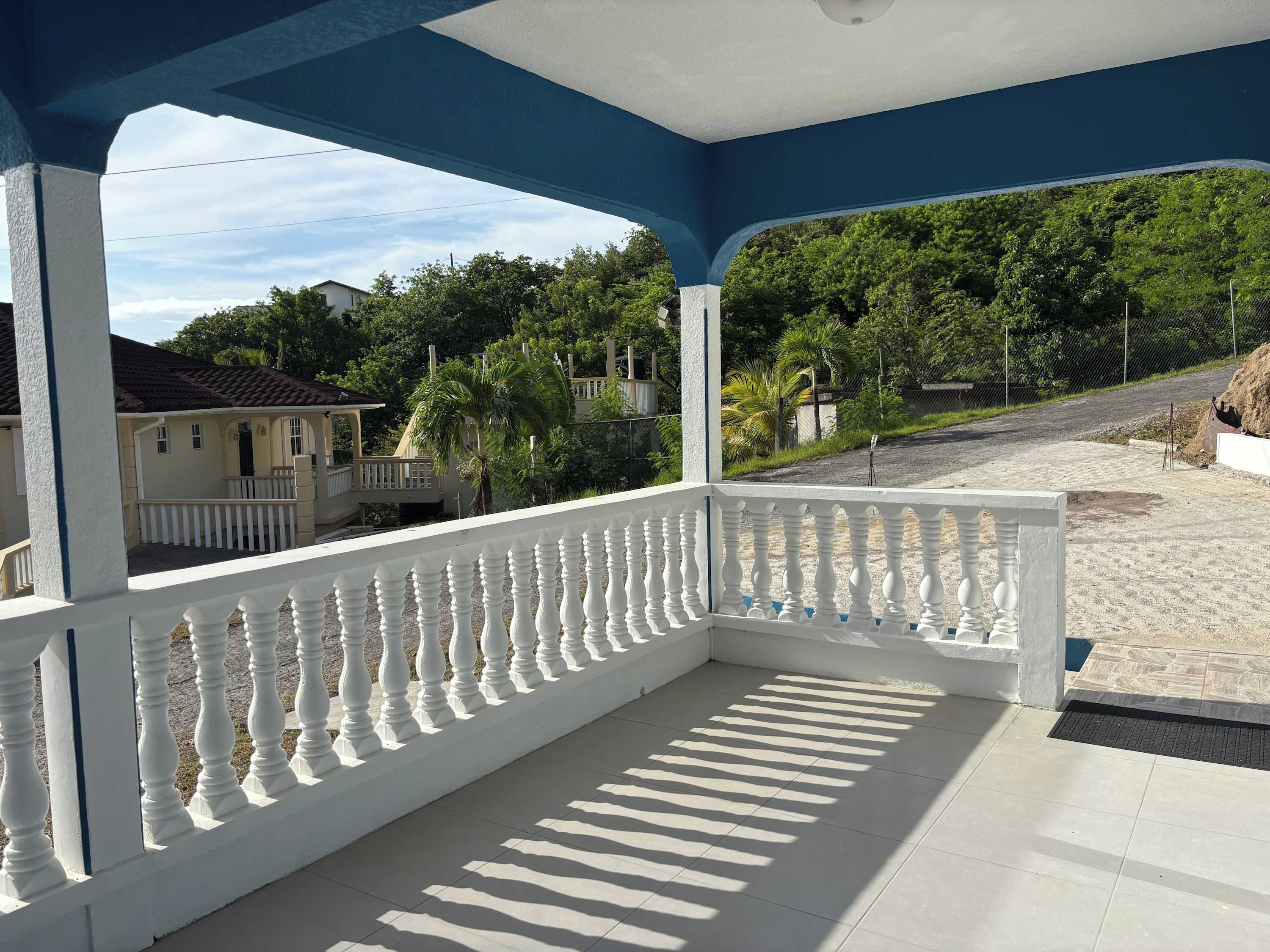 Un espacio exterior cubierto con balaustrada blanca, piso de baldosas y vista a una calle residencial con casas y vegetación.