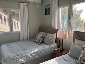 Ein Schlafzimmer mit Bett, Nachttisch, Lampe und Blick auf eine Treppe und Grünfläche vor dem Fenster.
