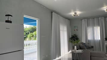 A modern living room with a grey sofa, a dining table, and a refrigerator. There is a balcony with white railings and a view of greenery.