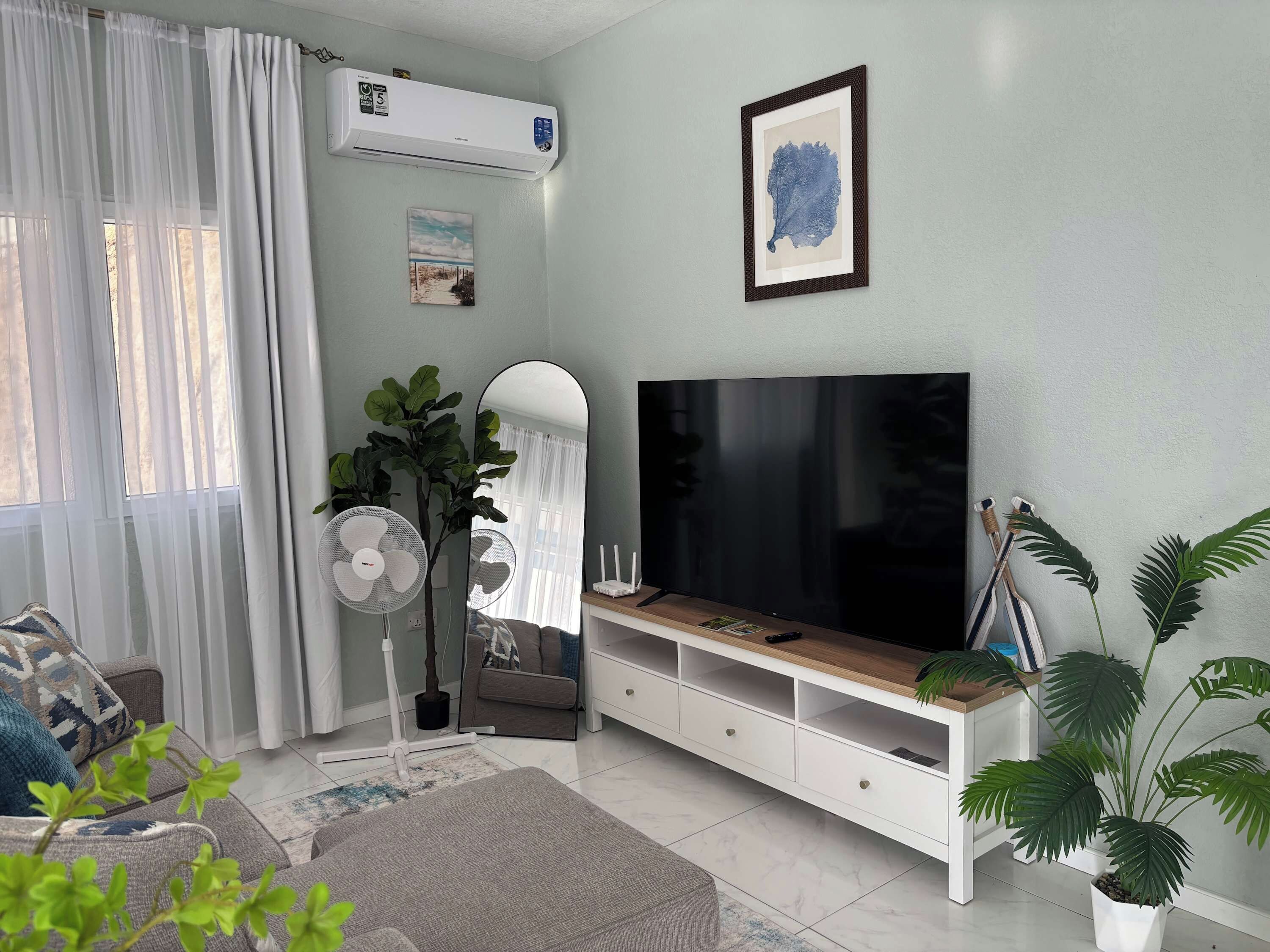 A living room with a flat-screen TV, a white entertainment unit, a large potted plant, a standing fan, and a window with sheer curtains.