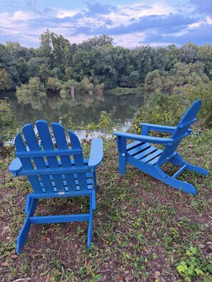 Two blue Adirondack chairs facing a calm lake surrounded by dense greenery.