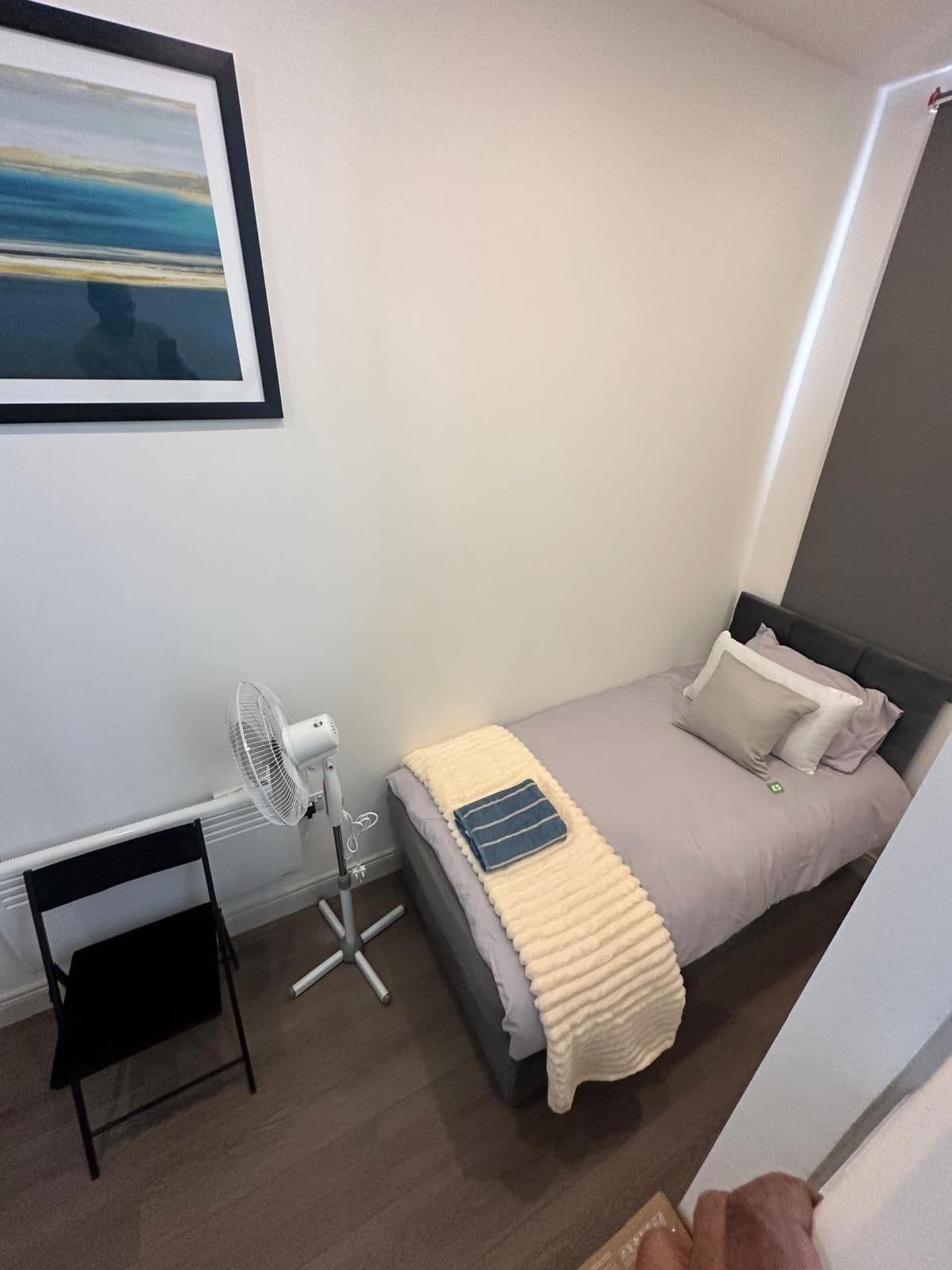 Een slaapkamer met een bed, een nachtkastje, een stoel, een ventilator en een ingelijste foto aan de muur.