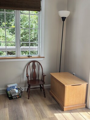 Ein Zimmer mit Fenster, einem Stuhl, einem Nachttisch und einer Lampe.
