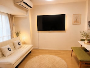 Een woonkamer met een bank, een flatscreen-televisie aan de muur, een rond tapijt en een eettafel met een bankje.