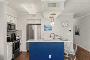 A modern kitchen with stainless steel appliances, a blue island, and a clock on the wall. - Destin West Pelican 308 - Amazing Views of the 700ft Lazy River & Emerald Waters! (Fort Walton Beach)