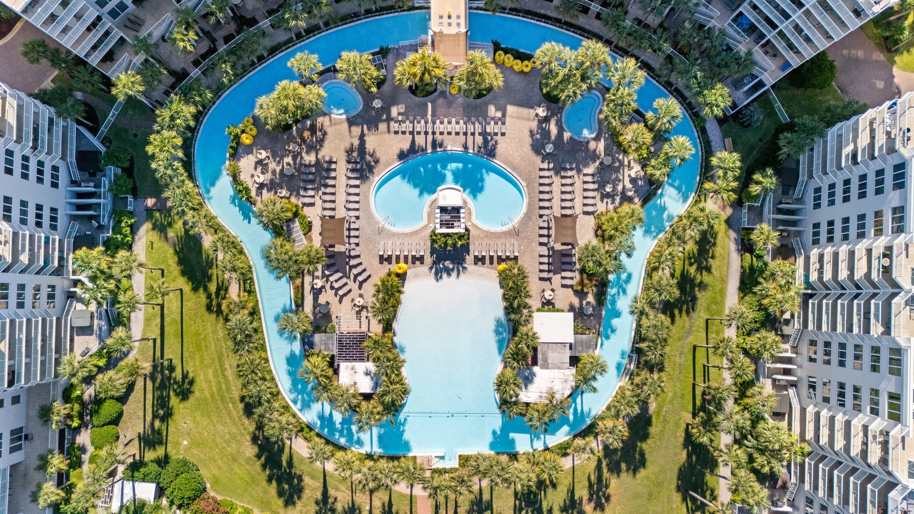 An aerial view of a hotel with a central swimming pool, multiple lounge areas, and surrounding greenery.