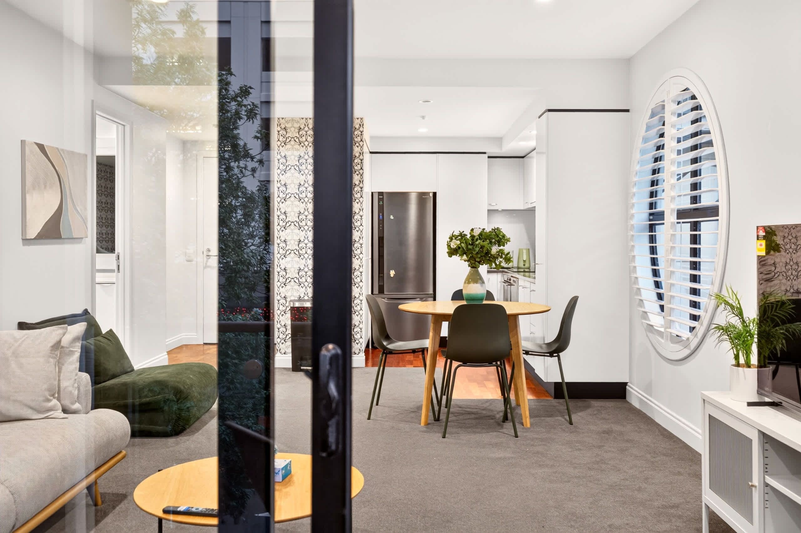 A modern living room with a sofa, a dining table with chairs, a kitchen area with a refrigerator, and a large window with blinds.