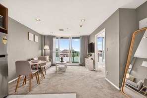 A modern living room with a sofa, dining table, and chairs, a flat-screen TV, and a view of a cityscape through large windows. - CBD Balcony Stay near Spark Arena | Zodiak Stays (Auckland)