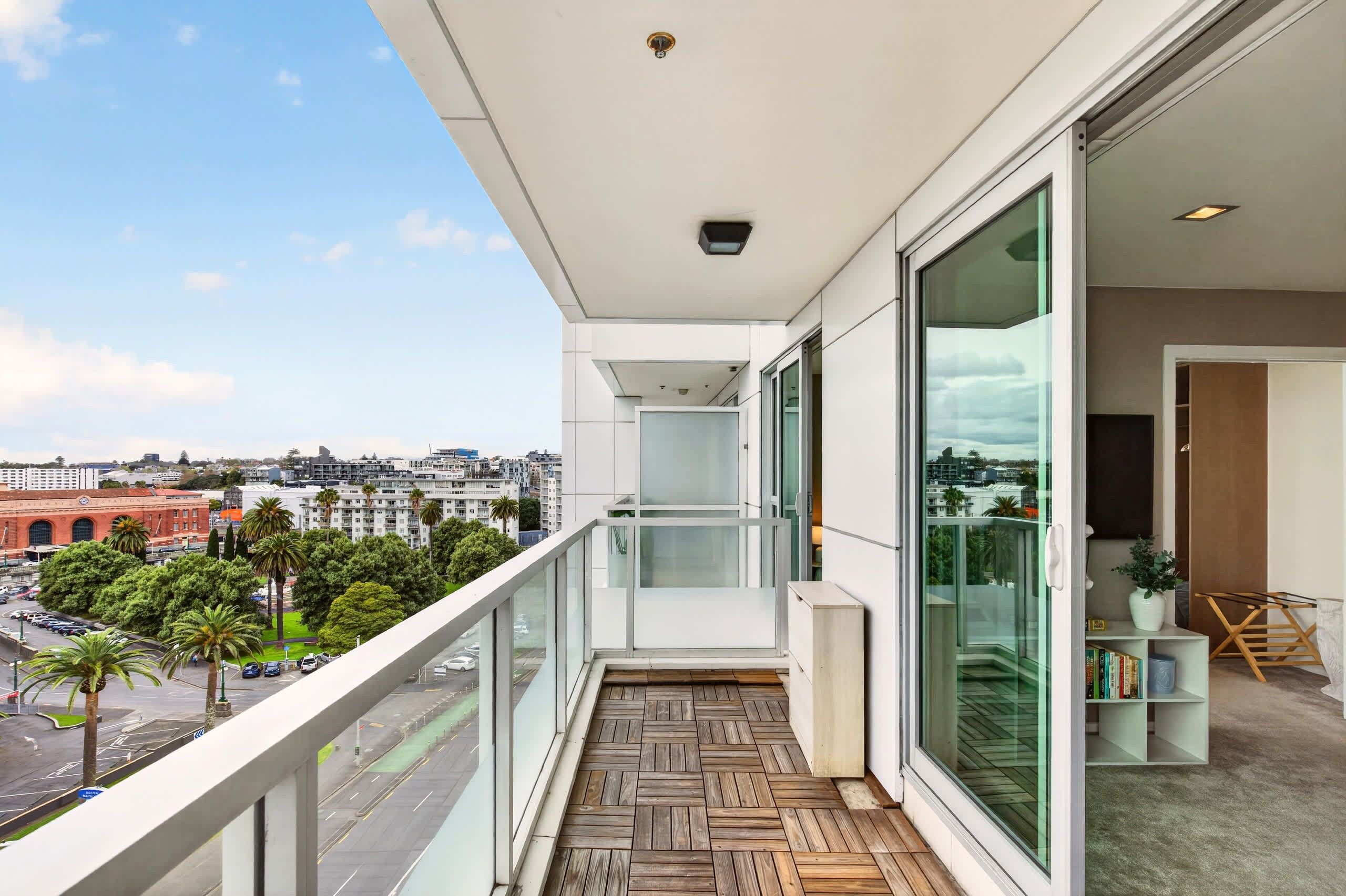 Un balcon donnant sur un paysage urbain, un plancher en bois et une porte vitrée menant à un espace intérieur agrémenté d’une bibliothèque et d’une plante.