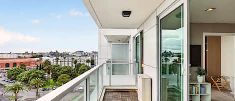 A balcony with a view of a cityscape, a wooden floor, and a glass door leading to an interior space with a bookshelf and a plant.