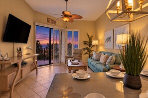 A living room with a TV, a ceiling fan, a sofa, a coffee table, and a view of the sunset through the balcony doors. - Phoenix East 110 | Beachside Outdoor Pool | Gulf Views | Pickleball Court (Orange Beach)