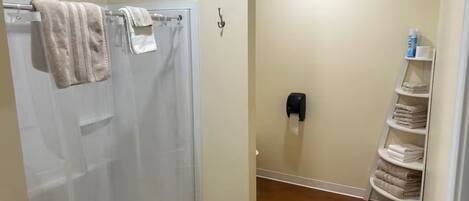 A bathroom with a shower, a towel rack, a soap dispenser, and a shelf with folded towels.