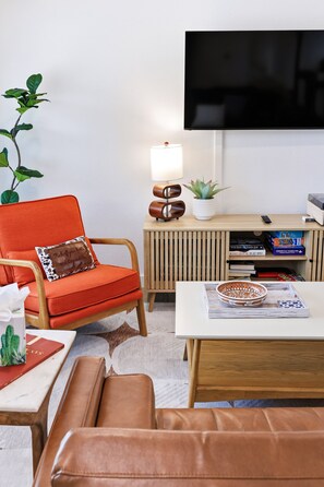 A living room with a TV mounted on the wall, a wooden side table with a lamp, a potted plant, and a bookshelf with books and games. - Agave - A Birdy Vacation Rental (San Antonio)