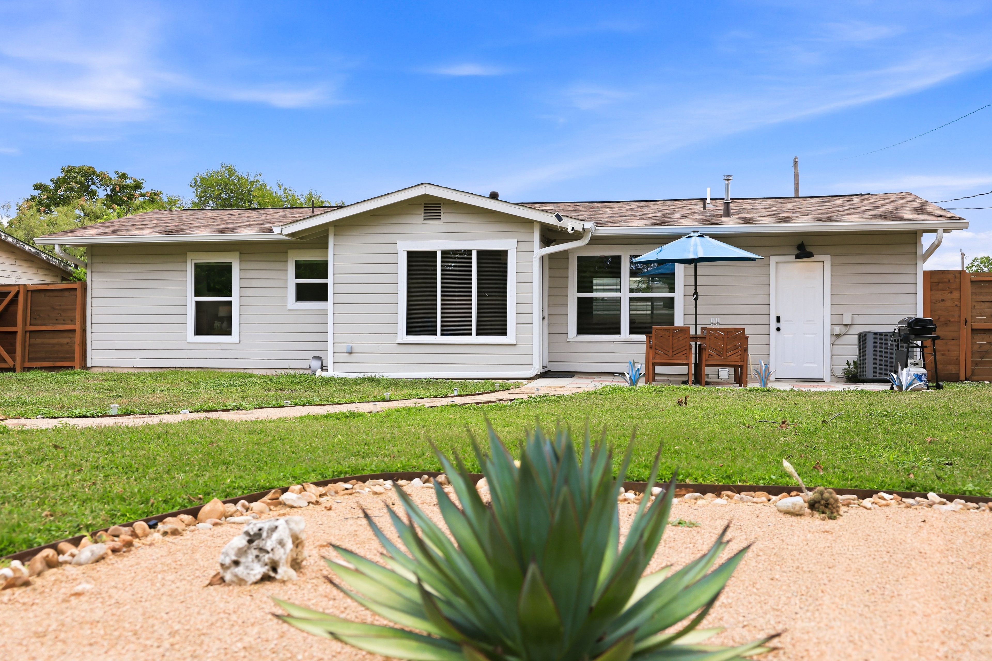 A single-story house with a front yard, a patio with a table and chairs, and a blue umbrella.