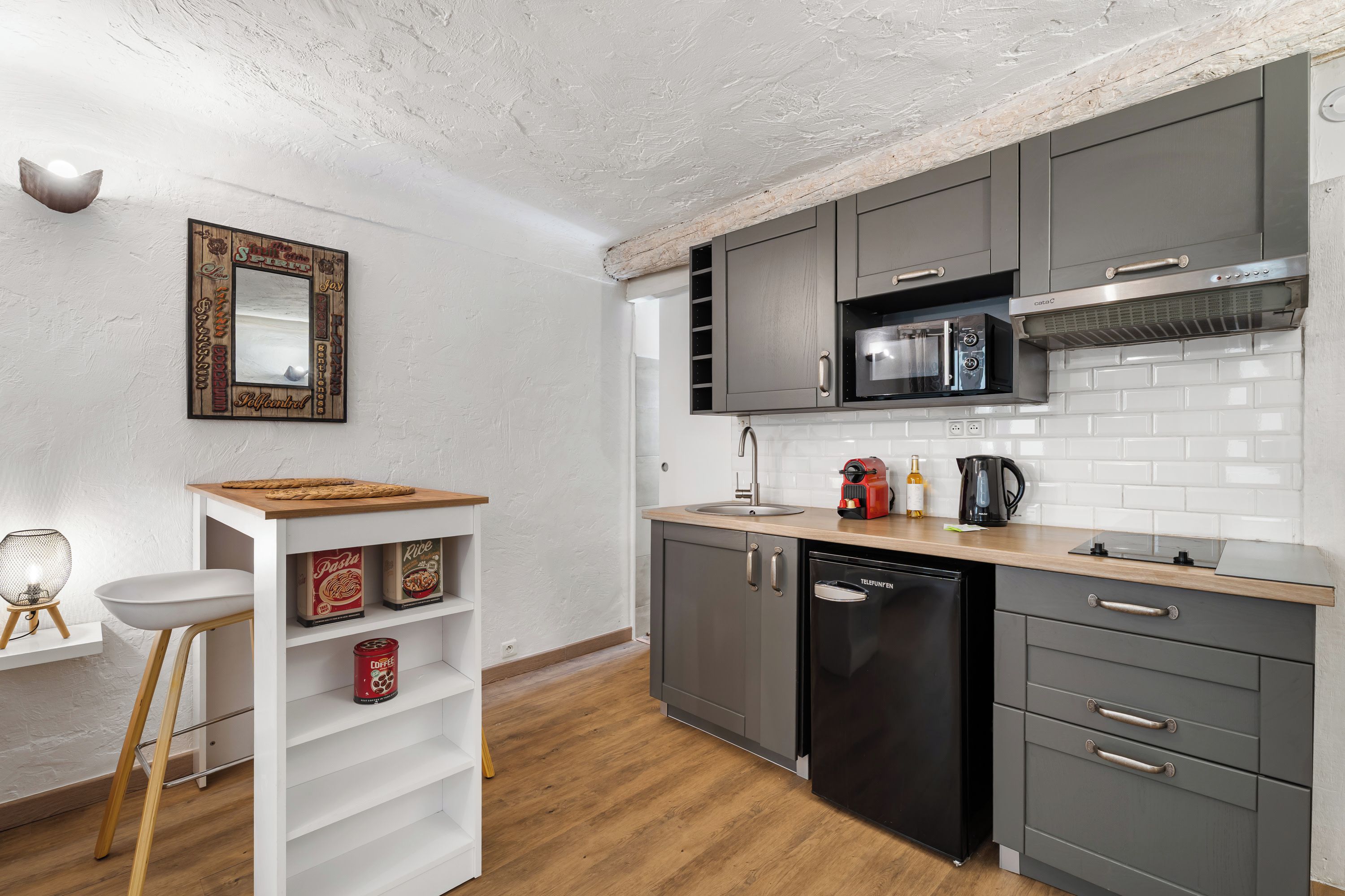 A modern kitchen with grey cabinets, a black dishwasher, and a microwave. There is a wooden stool with a woven seat and a white shelf with canned goods.