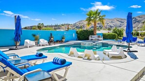 Un espace piscine avec des chaises longues et des parasols, un mur bleu et un paysage montagneux en arrière-plan.