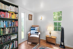 A room with a bookshelf, a chair, a lamp, a heater, and a window with a view of greenery. - Cozy Cottage Near Charlottesville & Wine Country (Charlottesville)