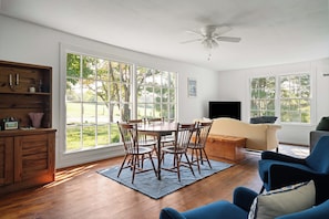 A spacious living room with a dining area, large windows, a ceiling fan, and a flat-screen TV.