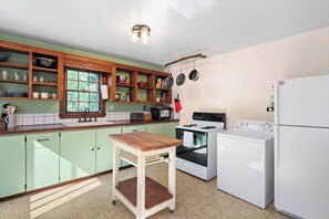 A kitchen with a wooden table, a stove, a refrigerator, and various kitchen utensils on shelves. - Cozy Cottage Near Charlottesville & Wine Country (Charlottesville)
