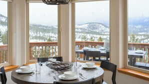 A dining area with a round table set for four, large windows offering a view of a snowy landscape, and a chandelier above. - Hilltop Haven with a Hot Tub, Game room, Poker lounge, Gym, Sauna, VIEWS (Bailey)
