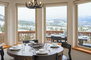Un coin repas avec une table ronde dressée pour quatre personnes, de grandes fenêtres donnant sur un paysage enneigé et un lustre au-dessus.