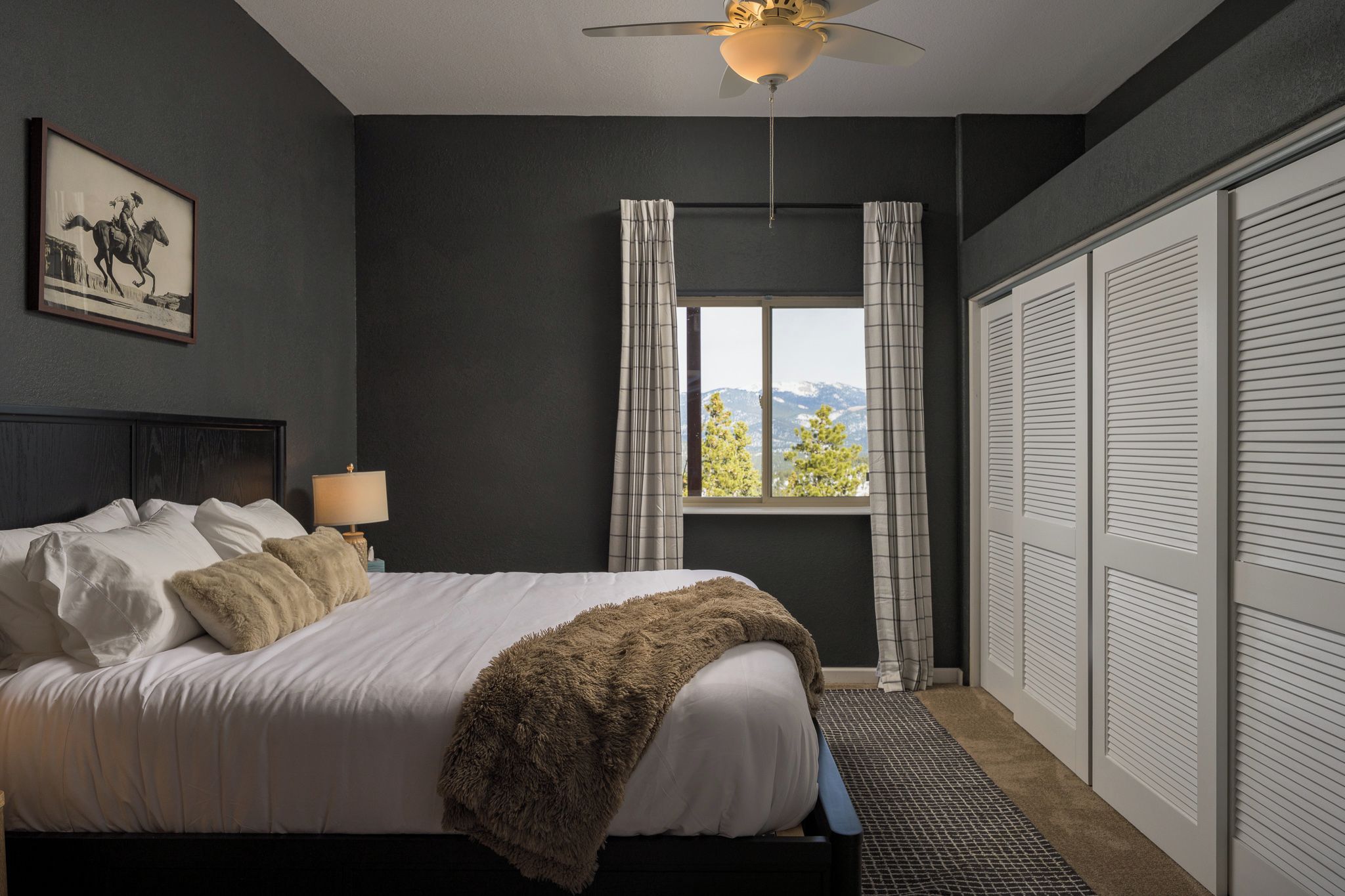 A bedroom with a bed, a nightstand, a window with curtains, and a view of mountains.