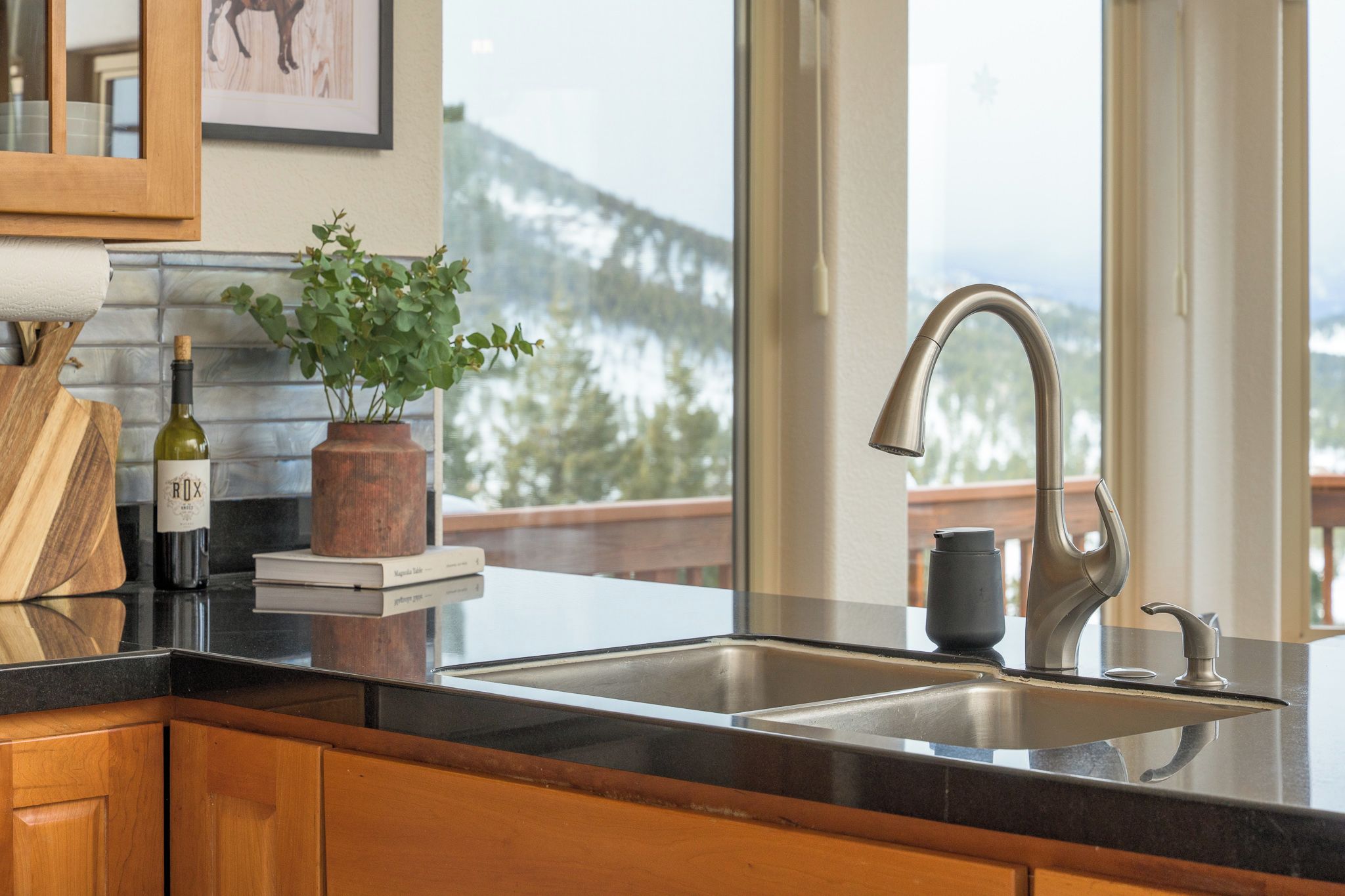 A kitchen with a stainless steel sink, wooden cabinets, a potted plant, a wine bottle, and a framed picture on the wall.