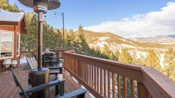 Una terraza de madera con un calentador metálico, mobiliario exterior y vistas a montañas nevadas.