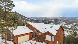 A wooden cabin with a garage, a deck, and a view of snow-covered mountains. - Hilltop Haven with a Hot Tub, Game room, Poker lounge, Gym, Sauna, VIEWS (Bailey)