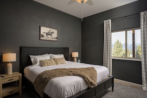 Une chambre à coucher comprenant un lit, deux tables de chevet, une commode, un ventilateur de plafond, une fenêtre avec des rideaux et un tableau encadré accroché au mur.