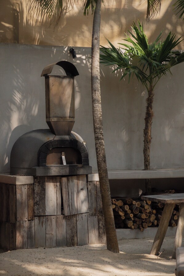 A rustic outdoor cooking area with a wood-fired oven, a wooden table, and a palm tree.