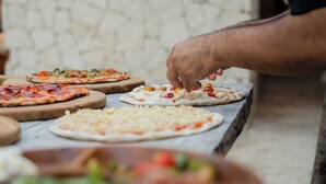 Uma pessoa preparando pizzas em uma bancada de mármore com diversos ingredientes e queijo.