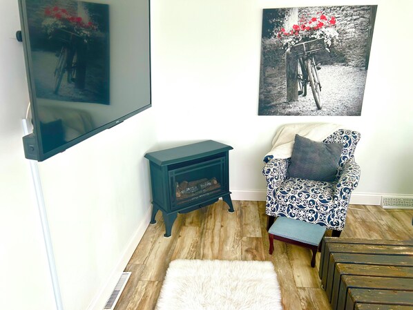 A living room with a television mounted on the wall, a small wooden table, a patterned armchair, a white furry rug, and a wooden bench.