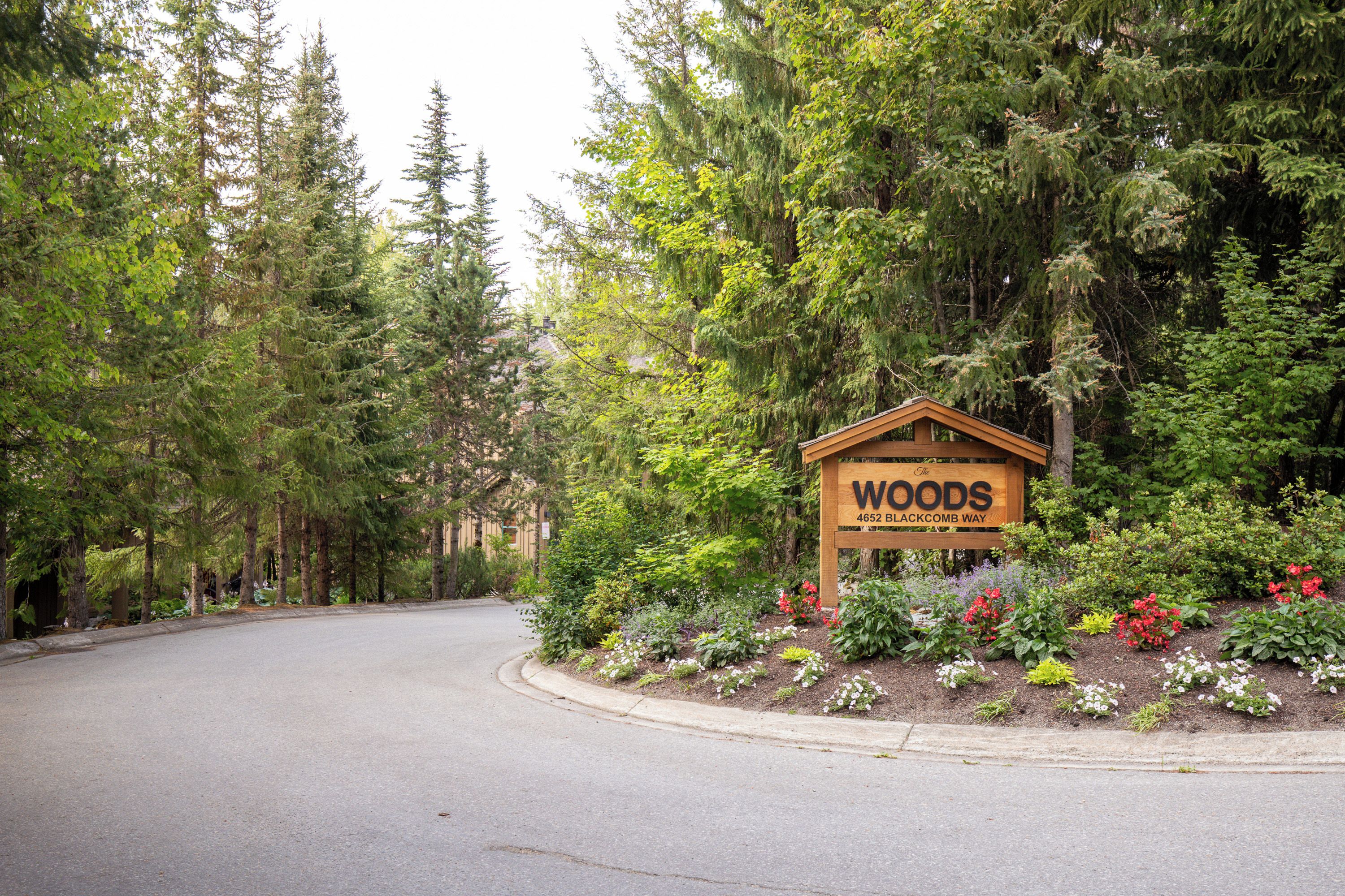 A sign for 'The Woods' at 4652 Blackcomb Way, surrounded by a forested area with a paved road.
