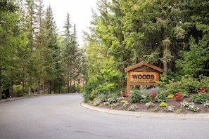 A sign for 'The Woods' at 4652 Blackcomb Way, surrounded by a forested area with a paved road. - Modern Mountain Retreat, Ski-In, Hot Tub (Whistler)