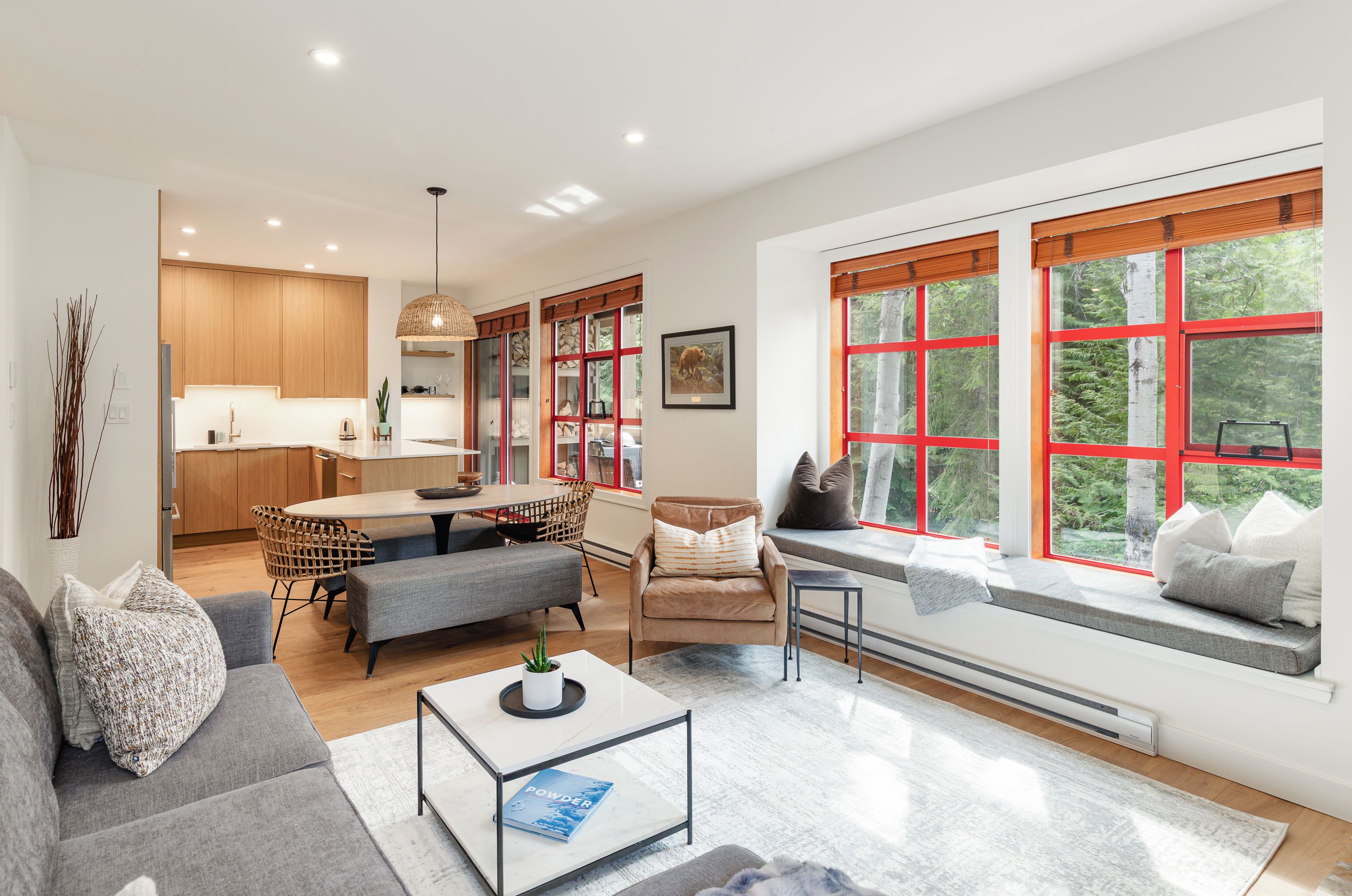 Una sala de estar moderna con sofá, mesa de centro y un asiento junto a la ventana con vista a árboles.