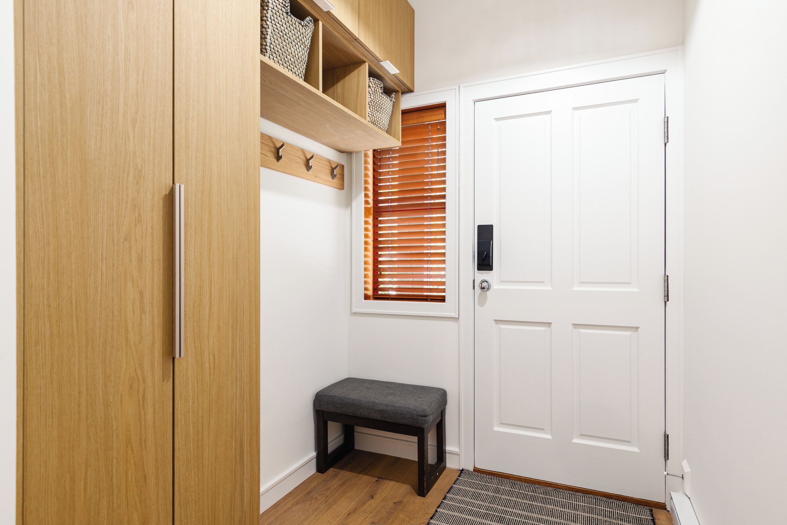 Un couloir avec une armoire en bois, un banc, une porte blanche et une fenêtre avec des stores.