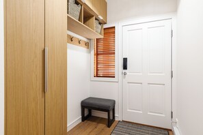Een gang met een houten garderobe, een bank, een witte deur en een raam met zonwering.