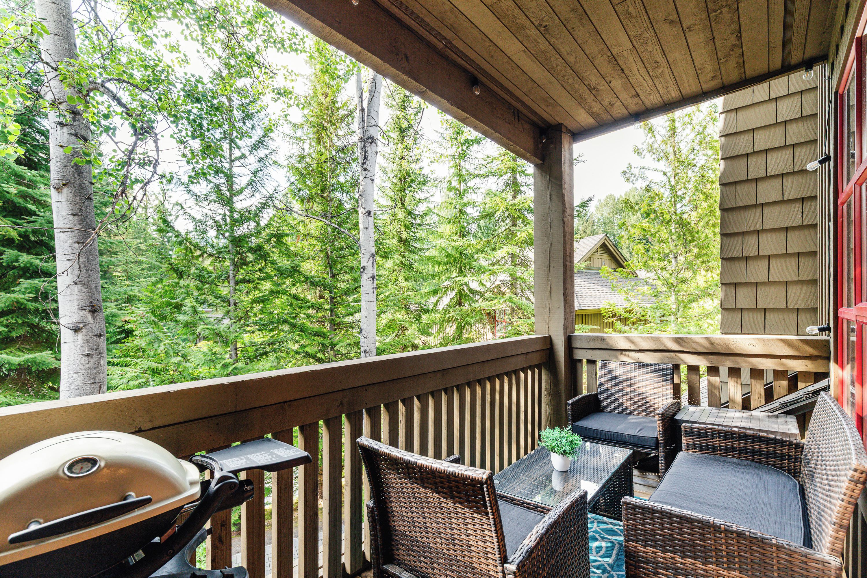 Ein überdachter Außenbereich mit Grill, Korbmöbeln und Blick auf einen Wald.