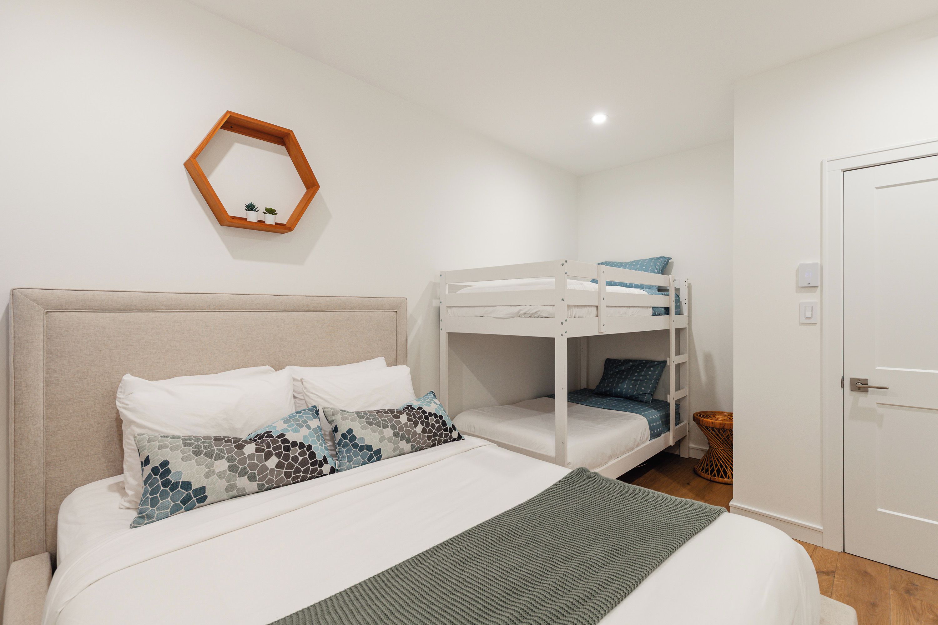 Un dormitorio con una cama litera, una cama individual con almohadas estampadas, un cabecero de madera, un espejo pequeño y una puerta.