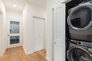 A laundry room with two stacked front-loading washing machines, a white door, and a wooden floor. - Modern Mountain Retreat, Ski-In, Hot Tub (Whistler)