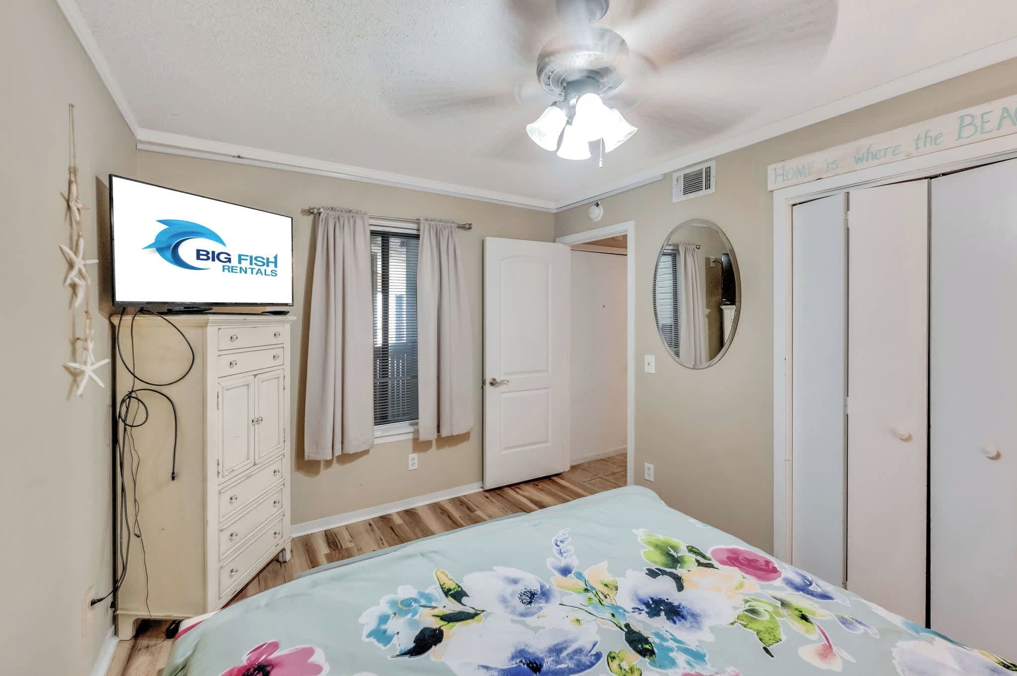 A bedroom with a bed, a ceiling fan, a TV, a dresser, a window with curtains, a mirror, and a door.