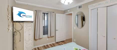 Un dormitorio con cama, ventilador de techo, televisor, cómoda, ventana con cortinas, espejo y puerta.